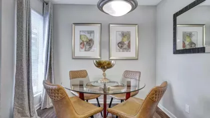 A cozy dining area with a round glass table, four tan upholstered chairs, and decorative wall art, illuminated by a modern pendant light.