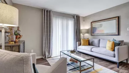 A bright and inviting living room with a gray sofa, accent chairs, a glass coffee table, and a large window with sliding doors and curtains.