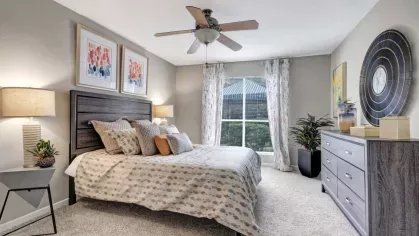 A spacious bedroom with a plush bed, patterned bedding, bedside lamps, and large windows, decorated with artwork and modern furniture.