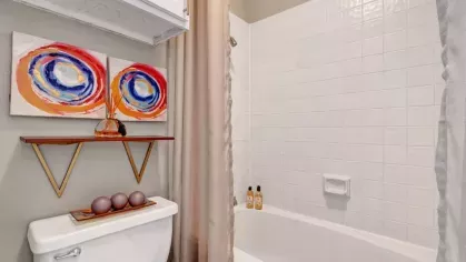 A bathtub with white tile surround, accented by colorful artwork, a wooden shelf, and a neutral shower curtain.