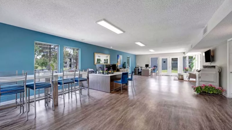 A spacious clubhouse with wood flooring, blue accents, and tables overlooking outdoor greenery.