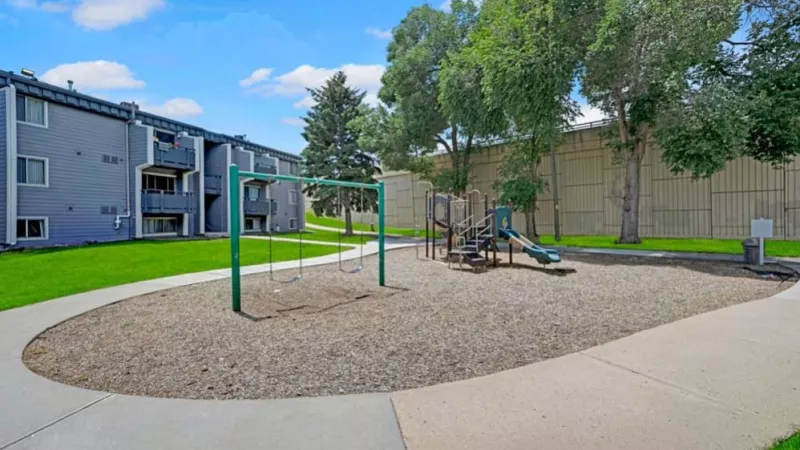 A playground area featuring swings, slides, and nearby apartment buildings shaded by tall trees.