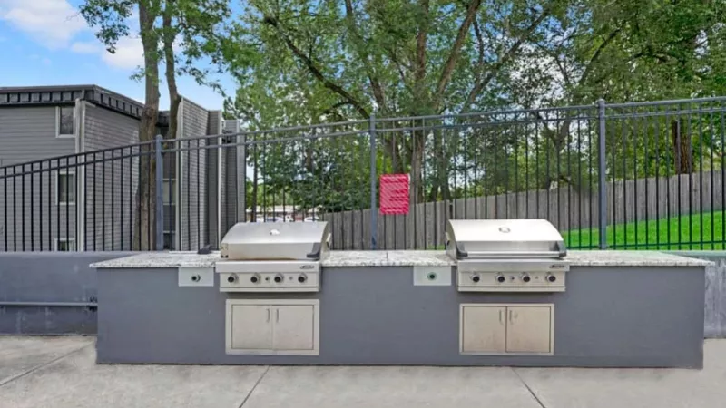 An outdoor grilling station with stainless steel barbecue units next to a fenced lawn area.