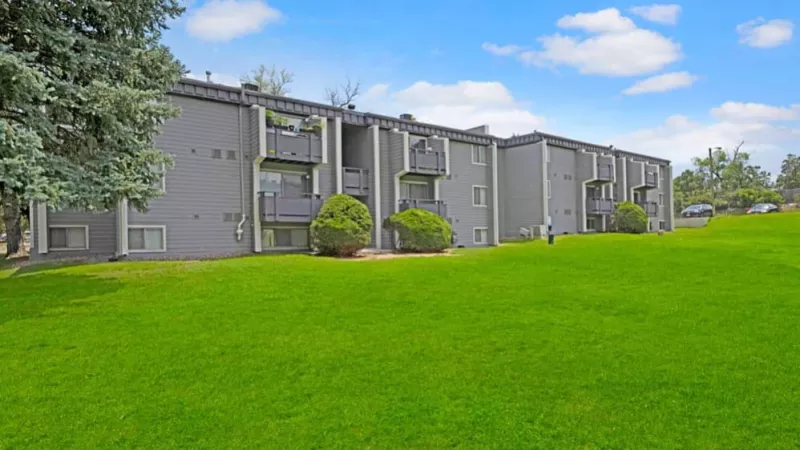 A broad stretch of green lawn in front of a gray apartment building with balconies and tall trees nearby.