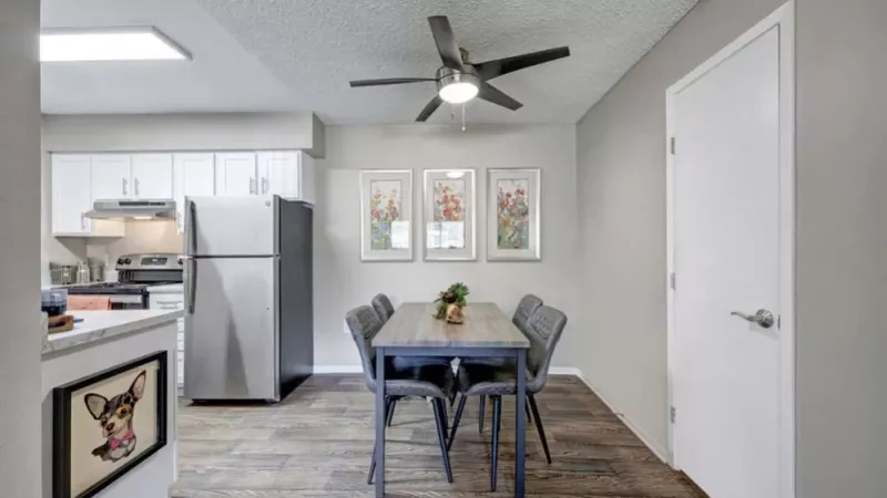 A cozy dining area with a modern table, chairs, and ceiling fan, adjoining a bright and functional kitchen.