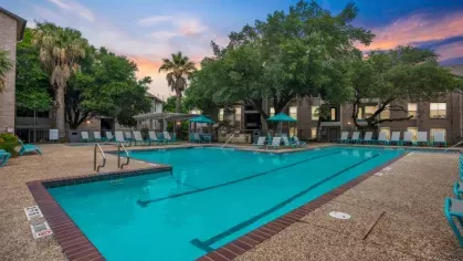 A large, rectangular swimming pool surrounded by lounge chairs and trees under a colorful sunset sky.