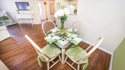 A dining area with a glass table set for four, featuring floral cushions and wooden flooring in a bright, open-concept living space.