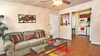 A cozy living room with a beige sofa, colorful throw pillows, a ceiling fan, and a view into the kitchen.