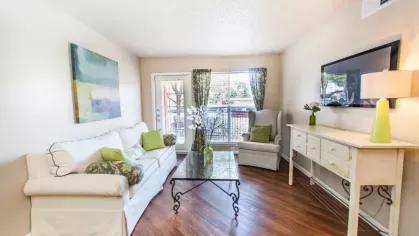 A living room with a white sofa, floral curtains, and green accents, featuring a glass coffee table and natural light streaming in.