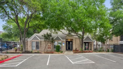 A hot tub adjacent to a swimming pool with shaded seating areas and tall trees in the background.