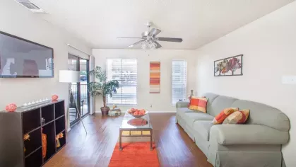 A vibrant living room with orange and yellow accents, a glass-top coffee table, and an open view into the kitchen.