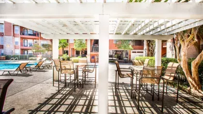 A shaded pergola area by the pool with outdoor seating and lounge chairs in a sunny apartment community courtyard.