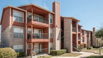 A three-story apartment building with red and beige exterior walls, surrounded by manicured bushes and trees.