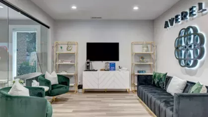 A stylish lounge with emerald green chairs, a plush velvet couch, gold-accented decor, and the Averelle North Hills logo on the wall.