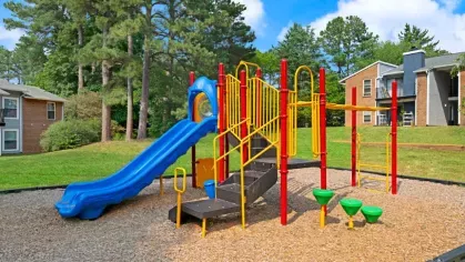 A colorful playground with climbing equipment, slides, and interactive features, set on a mulch surface with surrounding greenery.