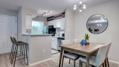 A stylish dining area with a wooden table, upholstered chairs, a round mirror, and a modern light fixture, adjacent to a sleek kitchen with a breakfast bar and barstools.