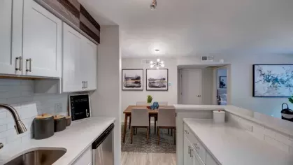 A contemporary kitchen with white cabinets, quartz countertops, stainless steel appliances, and a view into a dining area featuring modern artwork and lighting.