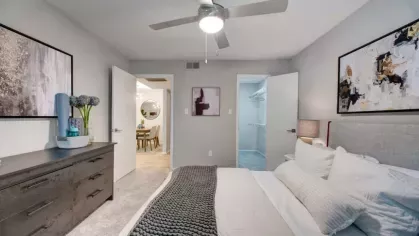 A serene bedroom with a ceiling fan, modern artwork, and a cozy bed, featuring a walk-in closet and neutral-toned decor.