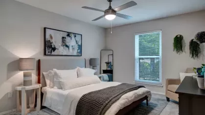 A bright bedroom with a large window, cozy bed, ceiling fan, modern artwork, and decorative plants, creating a calm and inviting space.