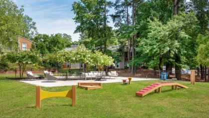 A fenced dog park with agility equipment and shaded seating areas surrounded by trees and greenery.