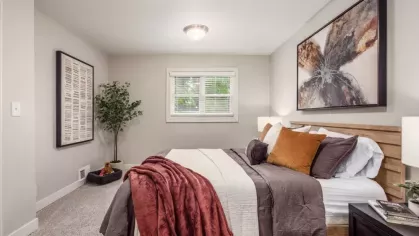 A second bedroom with a cozy setup, including warm-toned pillows and throws, a large window, and a wall-mounted coat hook with a backpack.