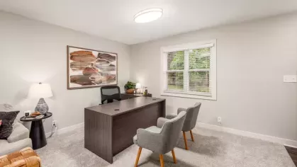 A professional home office environment with a large wooden desk, two comfortable chairs, and a large window providing ample natural light.