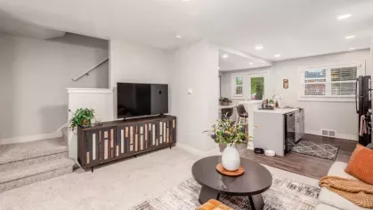 A living area with a flat-screen TV on a wooden console, a staircase leading upstairs, and an open-plan kitchen in the background.