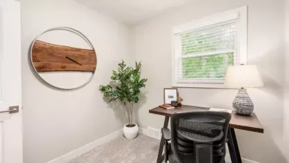 A minimalist home office with a large circular wooden wall clock, a desk, and a potted plant by the window.