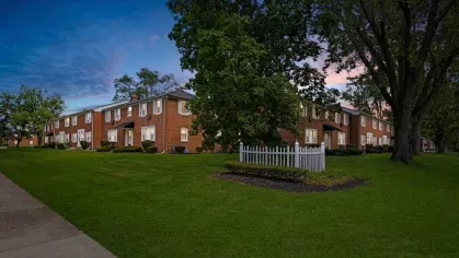 A brick apartment complex surrounded by lush green lawns and trees at twilight with a picket fence in the foreground.