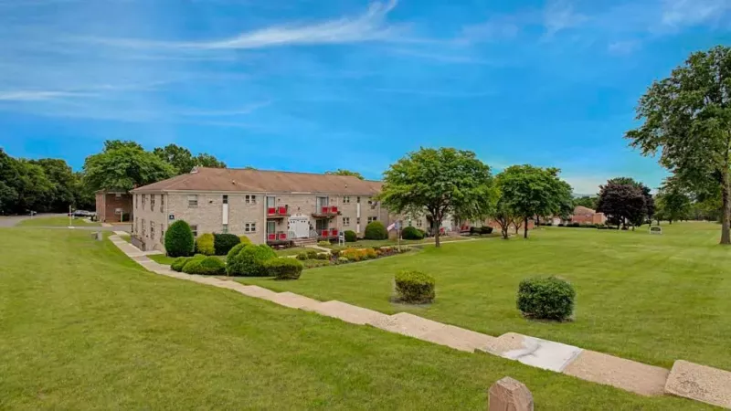 A green sign for "Brakeley Gardens" surrounded by well-maintained grass and shrubbery with apartment buildings in the background.