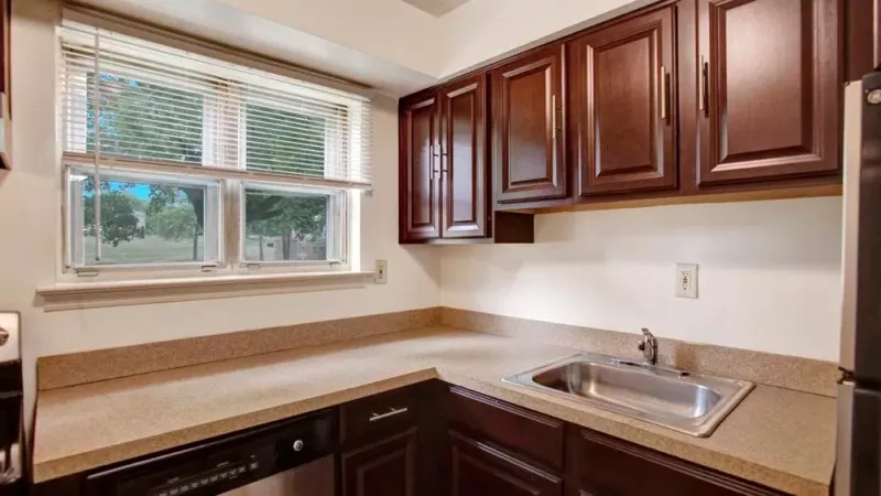 An open floor plan showcasing a living room with gray carpet, sliding glass doors leading to a balcony, and a kitchen with dark brown cabinets and stainless steel appliances.