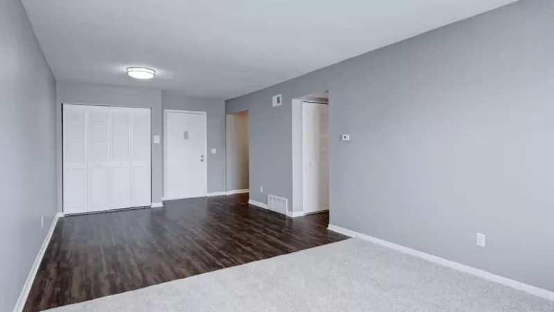 A spacious living room with a blend of hardwood flooring and carpet, featuring a large window and a closet with white doors.