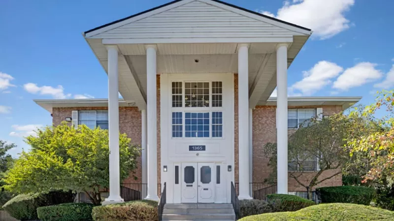 The front view of a two-story brick apartment building with a central entrance, white pillars, and a staircase leading up to the door.