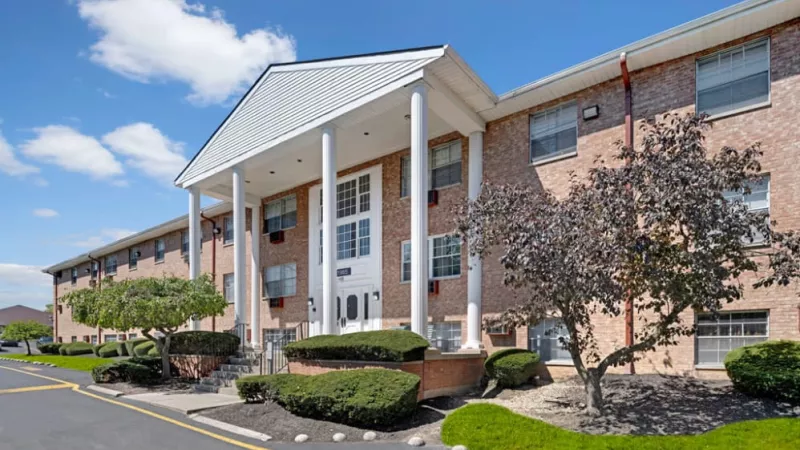 A side-angle view of the brick apartment building with white pillars, trimmed bushes, and a paved driveway.