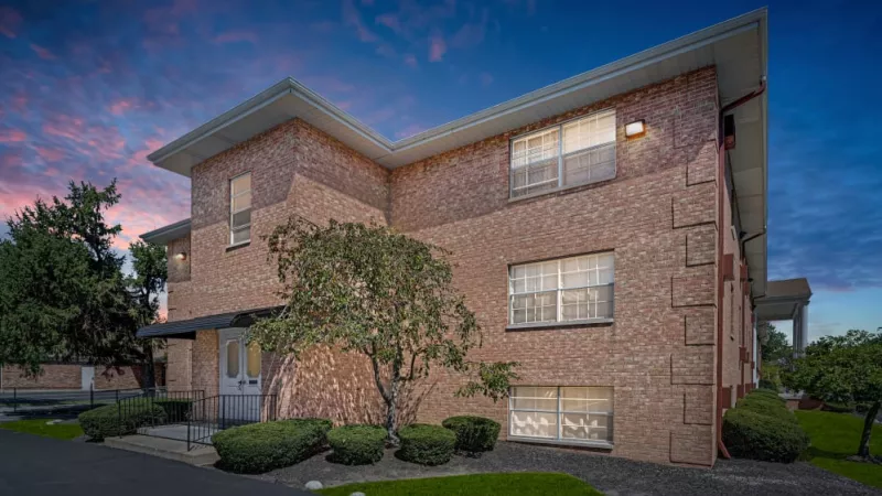 A corner perspective of a brick apartment building featuring a small tree and trimmed hedges during twilight.