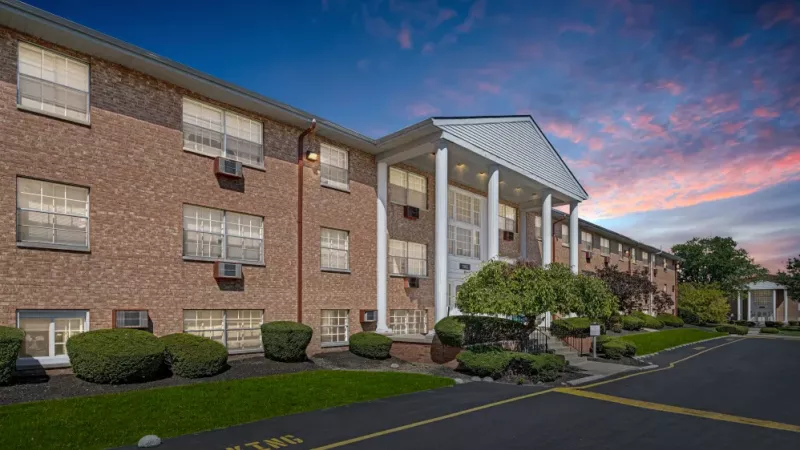 A wide-angle view of an apartment complex with well-maintained landscaping and a warm glow from the building lights at dusk.