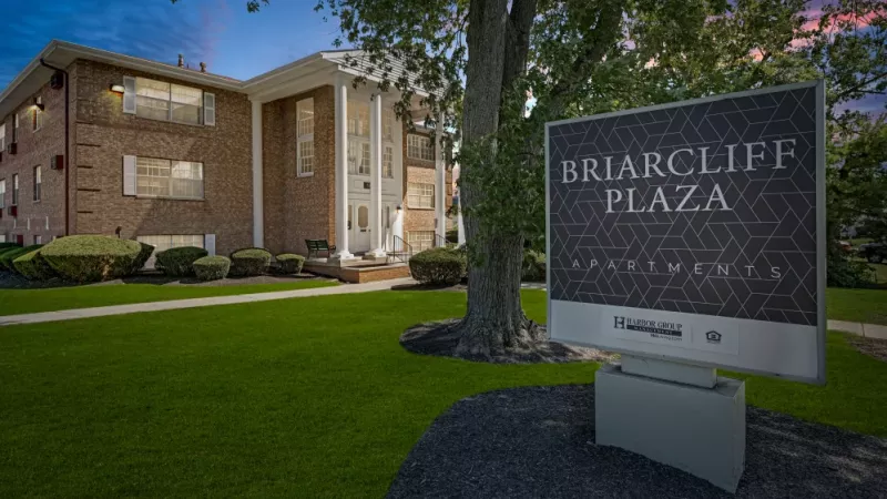 A twilight view of the Briarcliff Plaza Apartments sign and entrance, framed by trees and a beautifully manicured lawn.