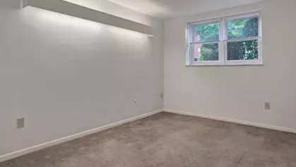 A bedroom with a carpeted floor, a window letting in natural light, and a white wall-mounted shelf light fixture.