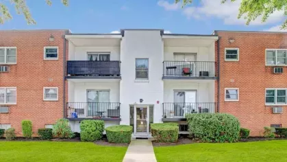 A front-facing view of a brick apartment building with balconies and neatly trimmed hedges.