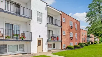 A side perspective of a white and brick apartment building featuring multiple balconies and green lawns.