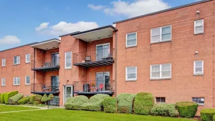 A side perspective of a red brick apartment complex with outdoor balconies and surrounding greenery.