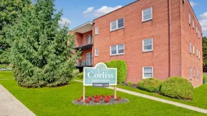 A red brick building with a sign that reads "Corliss Apartments" next to a large pine tree.