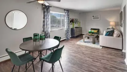 A spacious dining and living area with a round table, green chairs, and large windows.