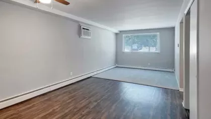 A bright living room with a mix of wood flooring and carpet, featuring a ceiling fan and large window.
