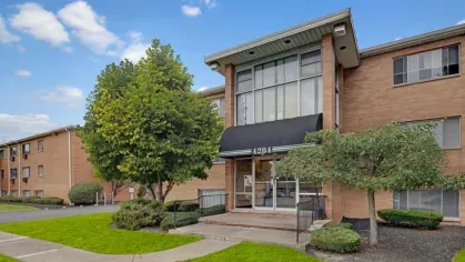 A closer view of the apartment building entrance with a black awning and manicured shrubs.