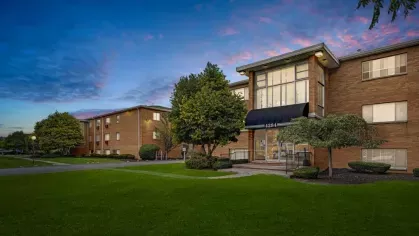 The facade of a brick apartment building at dusk, featuring large windows and lush greenery.