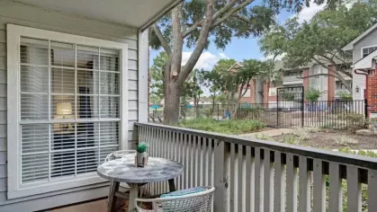 A private patio with a small round table and two chairs, overlooking a landscaped courtyard with trees and a pool in the distance.