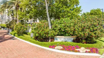 A landscaped entrance to The Heritage at Boca Raton featuring vibrant flowers, greenery, and a welcoming sign under palm trees.