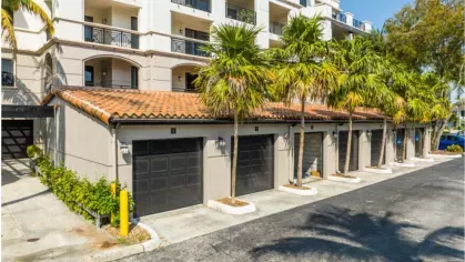 A row of garage units with terracotta roofs, palm trees, and part of the building's exterior with balconies in the background.