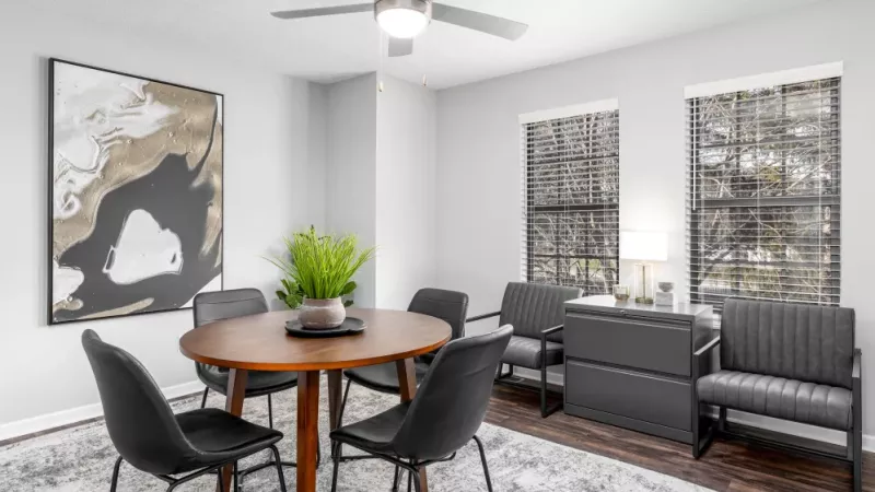 A dining area with a round wooden table, black chairs, and large windows letting in natural light.